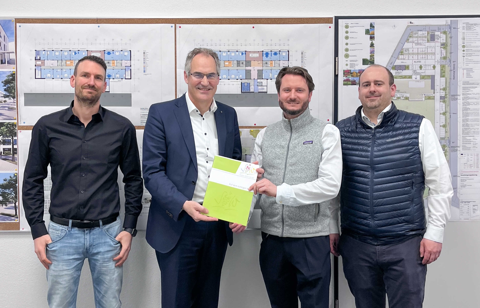 Vier Personen stehen vor einer Pinnwand mit Bildern und Plänen. Die beiden Personen in der Mitte halten gemeinsam eine Mappe mit dem Logo des Landkreises Südliche Weinstraße.