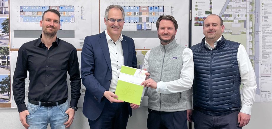 Vier Personen stehen vor einer Pinnwand mit Bildern und Plänen. Die beiden Personen in der Mitte halten gemeinsam eine Mappe mit dem Logo des Landkreises Südliche Weinstraße.