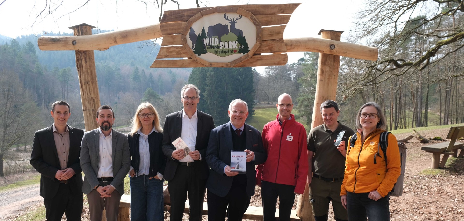 Neun Personen stehen unter einem hölzernen Eingangstor mit der Aufschrift „Wild- und Wanderpark“. Einige halten Flyer oder ein Smartphone in der Hand, im Hintergrund sind Wald und eine Wiese zu sehen.