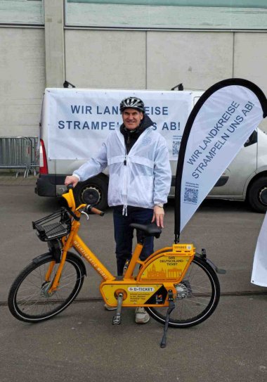 Dietmat Seefeldt neben gelbem Fahrrad und einer Beachflag mit Aufschrift "Wir Landkreise strampeln uns ab". Auf einem Kleinbus im Hintergrund ein Banner mit gleicher Aufschrift.