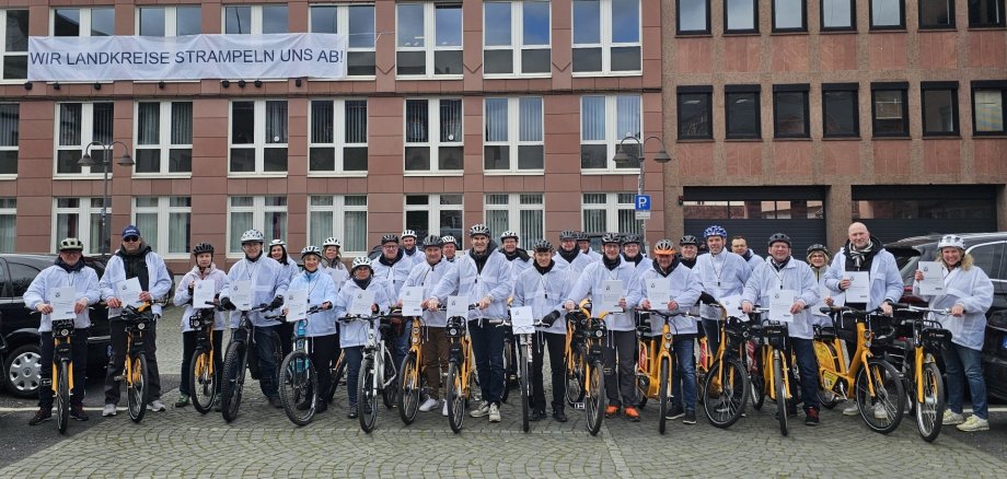 Gruppenbild vor Gebäude mit Banner mit der Aufschrift "Wir Landkreise strampeln uns ab". Alle tragen weiße Jacken.