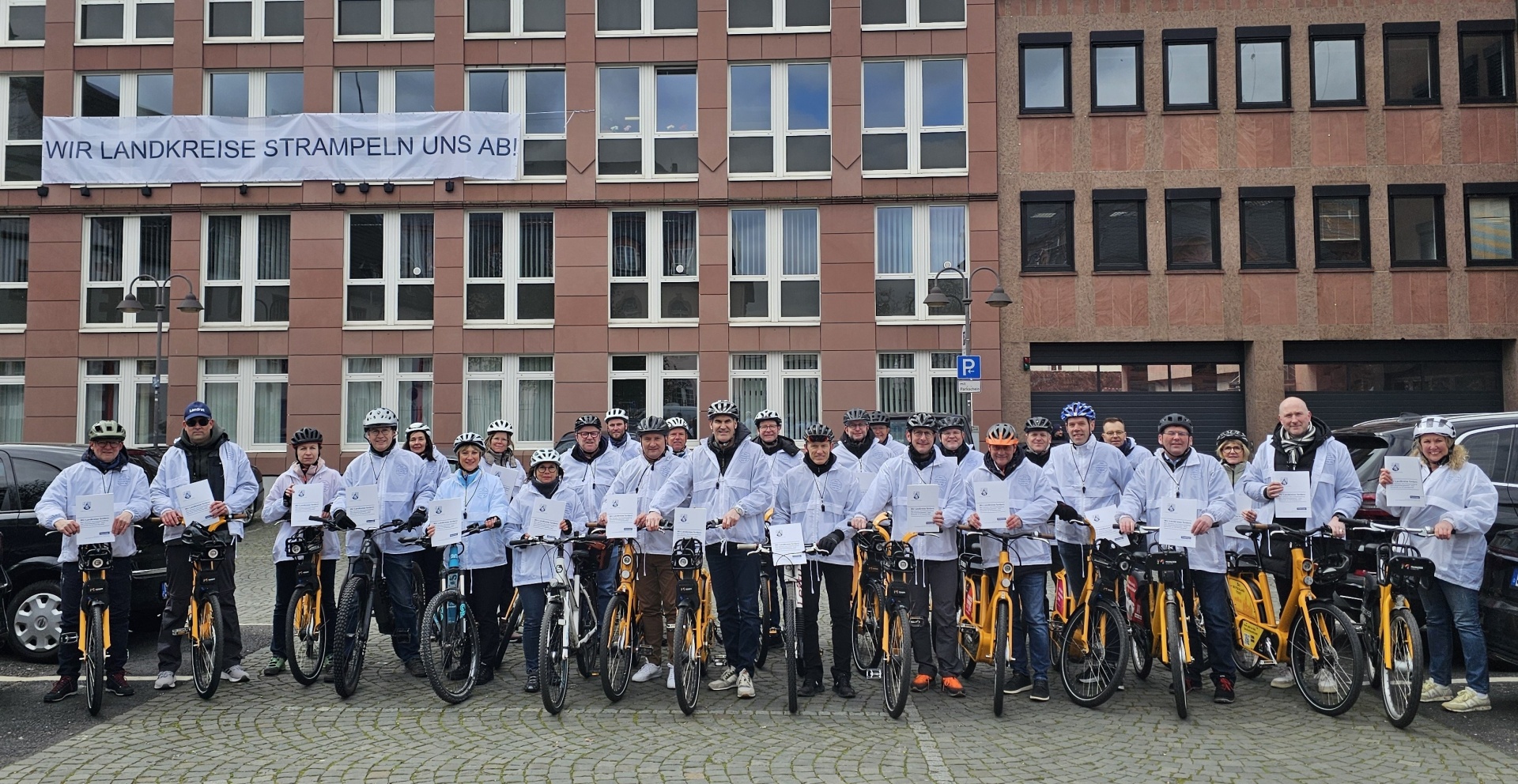 Gruppenbild vor Gebäude mit Banner mit der Aufschrift "Wir Landkreise strampeln uns ab". Alle tragen weiße Jacken.