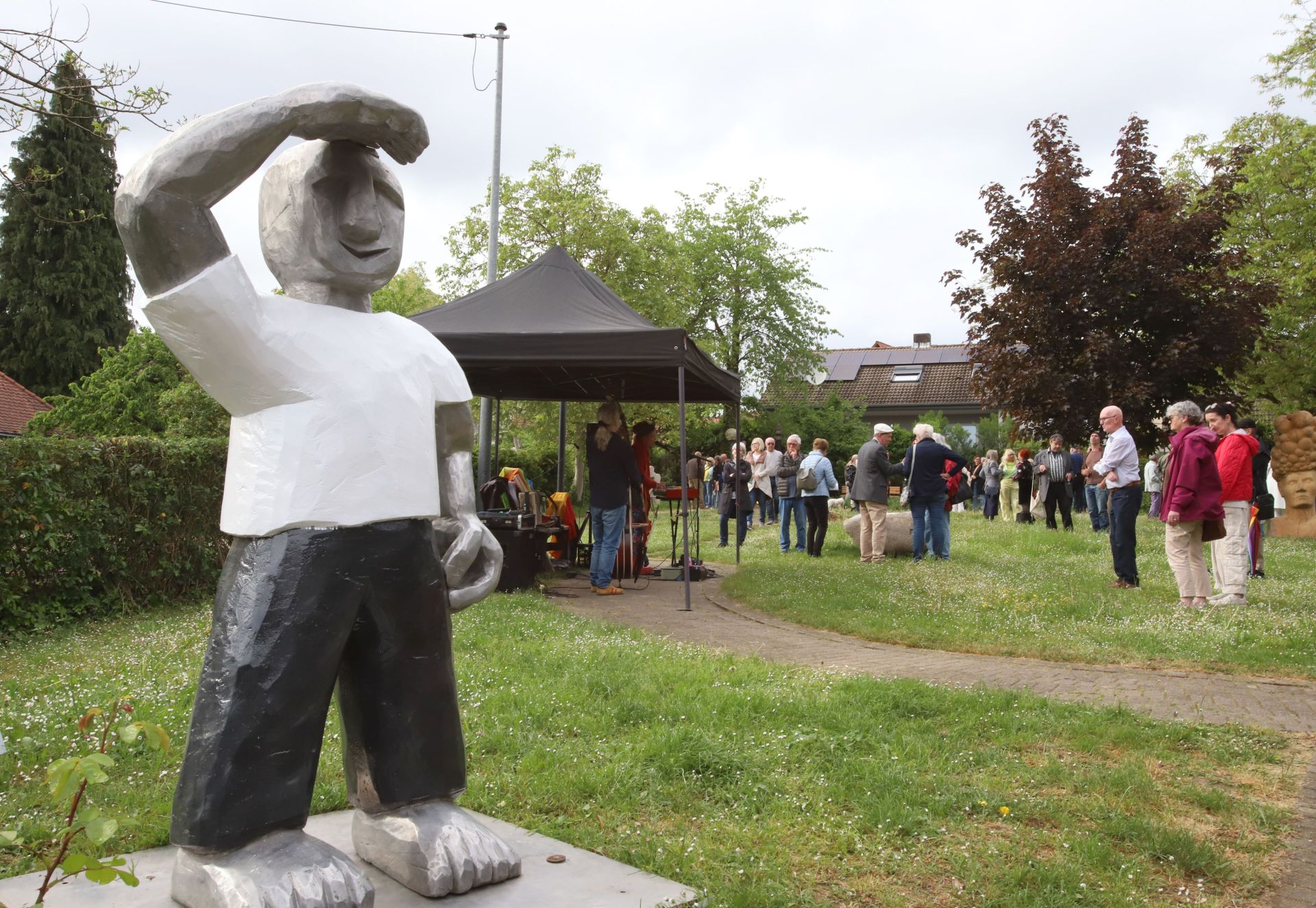 Im Vordergrund steht eine große Skulptur einer Figur, die eine Hand an die Stirn hält, als ob sie nach etwas Ausschau hält und Sonnenlicht abschirmt. Im Hintergrund besuchen zahlreiche Menschen eine Veranstaltung im Grünen.