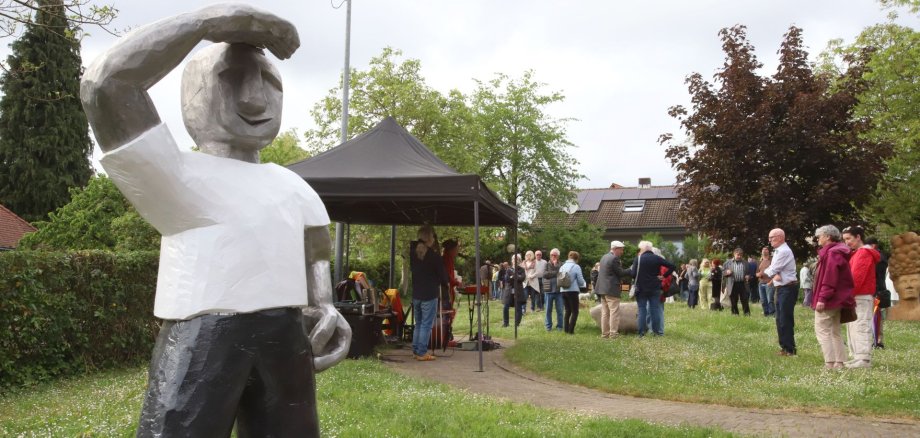 Der Skulpturengarten 2026 im Gemeindegarten Schweigen-Rechtenbach – hier bei der Eröffnung am 5. Mai – ist von Mai bis Oktober geöffnet. Foto: Björn Iversen/frei Im Vordergrund steht eine große Skulptur einer Figur, die eine Hand an die Stirn hält, als ob sie nach etwas Ausschau hält und Sonnenlicht abschirmt. Im Hintergrund besuchen zahlreiche Menschen eine Veranstaltung im Grünen.