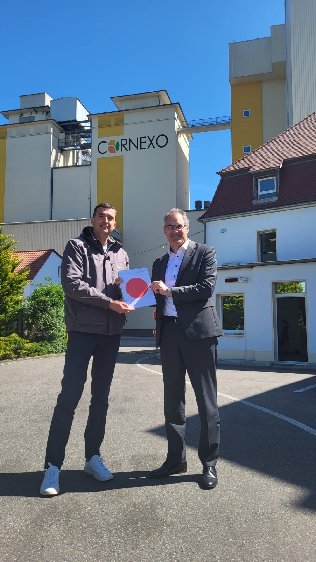 Zwei Männer stehen vor den Gebäuden des Unternehmens Cornexo und halten gemeinsam eine Baugenehmigung mit rotem Punkt in die Kamera. Im Hintergrund sind die Produktionsgebäude des Unternehmens unter blauem Himmel zu sehen.