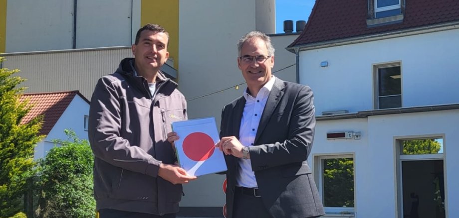 Landrat Dietmar Seefeldt (rechts) und Patrick Bindewald, geschäftsführender Gesellschafter der Cornexo GmbH, bei der Übergabe der Baugenehmigung. Foto: Cornexo Zwei Männer stehen vor den Gebäuden des Unternehmens Cornexo und halten gemeinsam eine Baugenehmigung mit rotem Punkt in die Kamera. Im Hintergrund sind die Produktionsgebäude des Unternehmens unter blauem Himmel zu sehen.