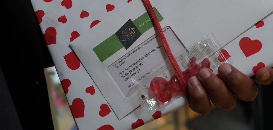 Nahaufnahme Geschenk und Umschlag mit Adresse einer Kita. Davor Fruchtgummi in "Danke"-Form.