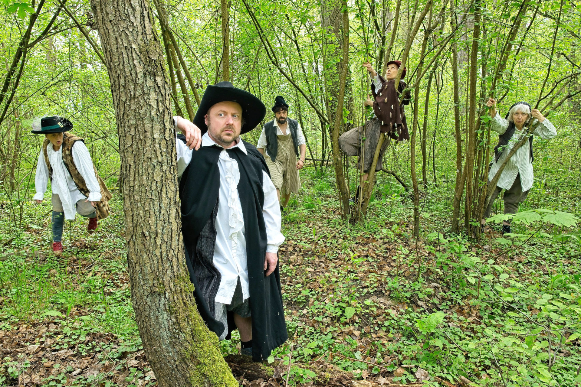 Fünf Personen in mittelalterlicher oder frühneuzeitlicher Kleidung stehen verteilt in einem Waldstück.