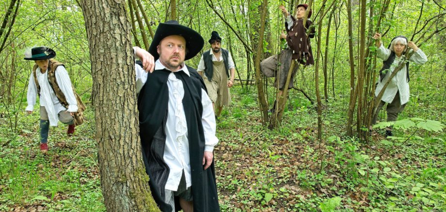 Fünf Personen in mittelalterlicher oder frühneuzeitlicher Kleidung stehen verteilt in einem Waldstück.