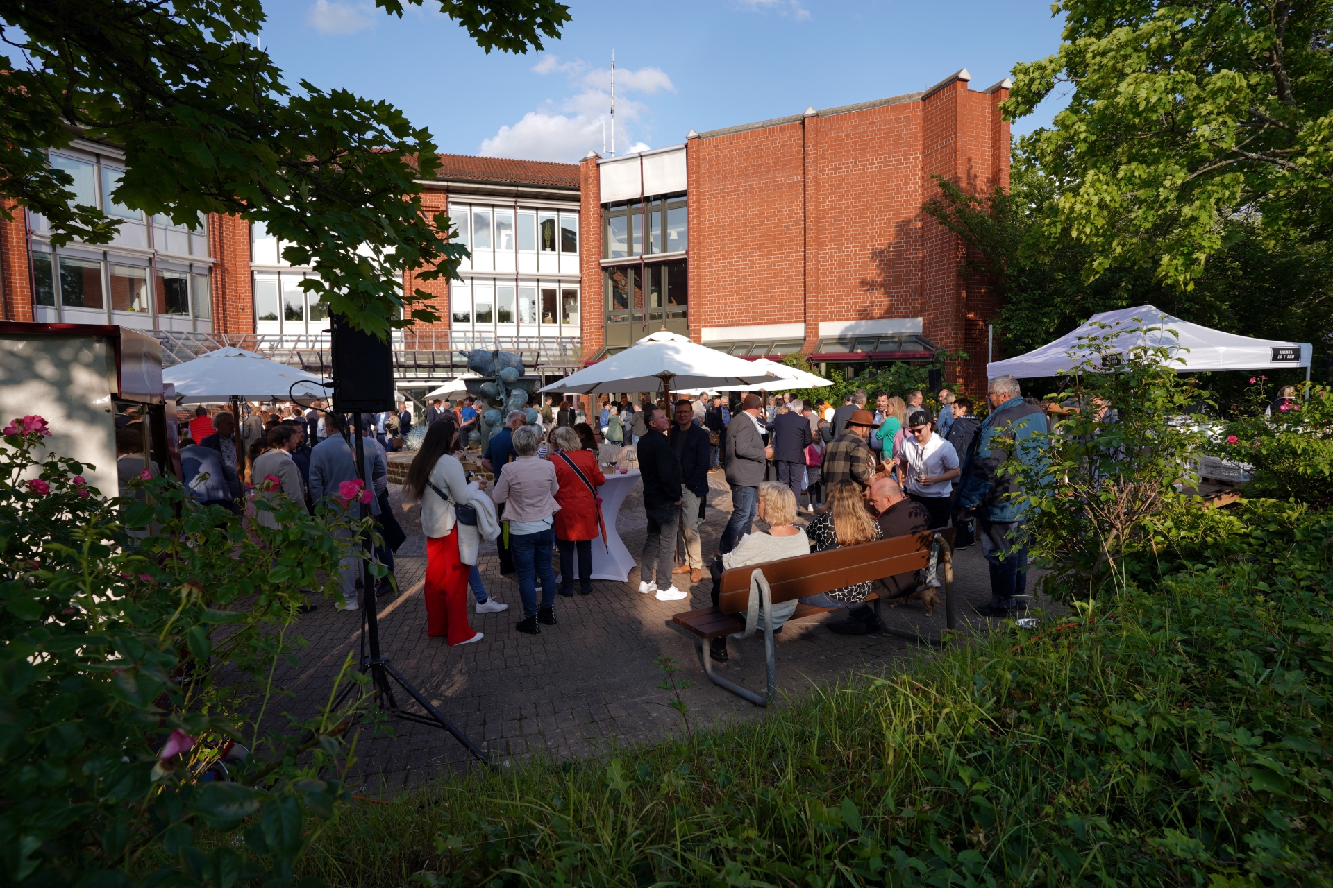 Eine Outdoor-Veranstaltung auf dem Platz vor dem Kreishaus der Kreisverwaltung Südliche Weinstraße. Menschen stehen unter weißen Sonnenschirmen vor dem roten Backsteingebäude der Kreisverwaltung. Das Wetter ist sonnig und die Veranstaltung ist gut besucht, mit einer vielfältigen Gruppe von Menschen, die sich unterhalten und das Event genießen.