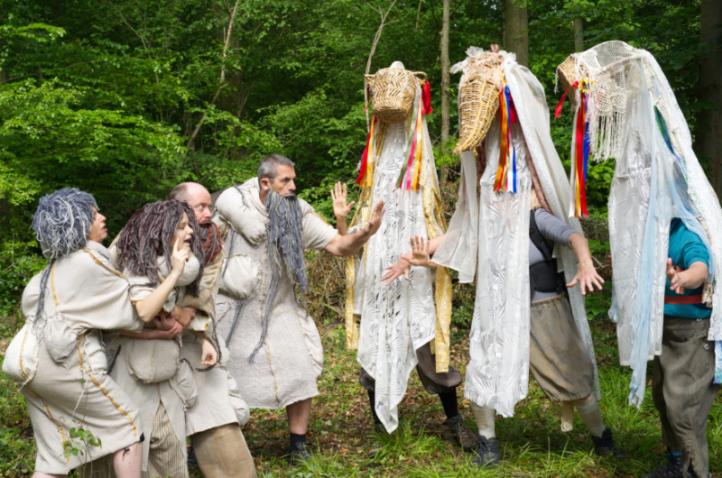 Schauspielszene im Wald: Mehrere kostümierte Personen in hellen Gewändern begegnen zwei hochgewachsenen Figuren mit maskenartigen Kopfbedeckungen. Die Szene wirkt märchenhaft und geheimnisvoll.