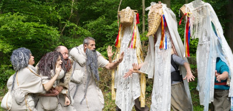 Schauspielszene im Wald: Mehrere kostümierte Personen in hellen Gewändern begegnen zwei hochgewachsenen Figuren mit maskenartigen Kopfbedeckungen. Die Szene wirkt märchenhaft und geheimnisvoll.
