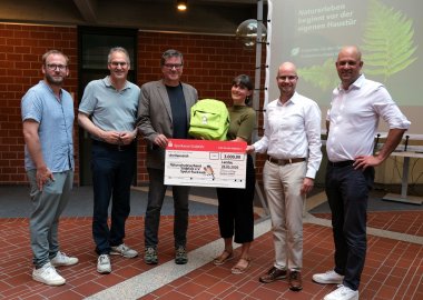 Bei der Vorstellung des „Spetzi Entdeckerrucksacks Wald“ im Kreishaus SÜW (von links): Beigeordneter der Verbandsgemeinde Edenkoben Daniel Poth in Vertretung für Bürgermeister Daniel Salm, Landrat Dietmar Seefeldt (SÜW), NVS-Vorsitzender Kurt Garrecht, Meike Wagner vom NVS, Svend Larsen, Vorstandsvorsitzender der Sparkasse Südpfalz, und Landrat Martin Brandl (GER). Foto: KV SÜW Sechs Personen präsentieren einen symbolischen Spendenscheck über 3.000 Euro. Eine Teilnehmerin hält den grünen „Spetzi Entdeckerrucksack Wald“ in die Kamera. Im Hintergrund ist eine Präsentationsfolie mit einem Farnblatt und dem Slogan „Naturerleben beginnt vor der eigenen Haustür“ zu sehen.