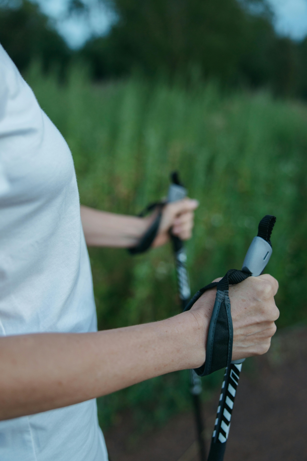 Person mit hellem T-Shirt hält beim Gehen zwei Nordic-Walking-Stöcke in den Händen. Im Hintergrund sind ein Weg und eine grüne, unscharf dargestellte Landschaft zu sehen.