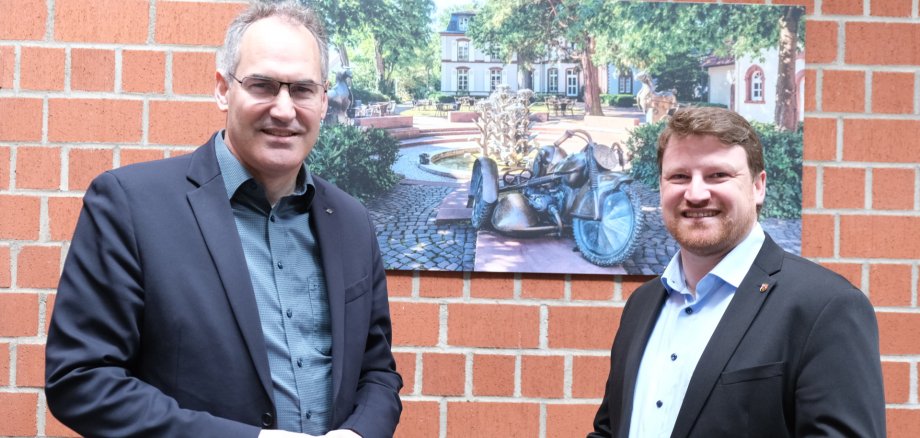 Landrat Dietmar Seefeldt (links) und Sven Koch im Jahr 2024 im Kreishaus SÜW, vor einem Foto des Herxheimer Dorfbrunnens. Archivfoto: KV SÜW Sven Koch und Landrat Dietmar Seefeldt stehen vor einem Bild im Kreishaus, das den Tabakbrunnen in Herxheim zeigt.