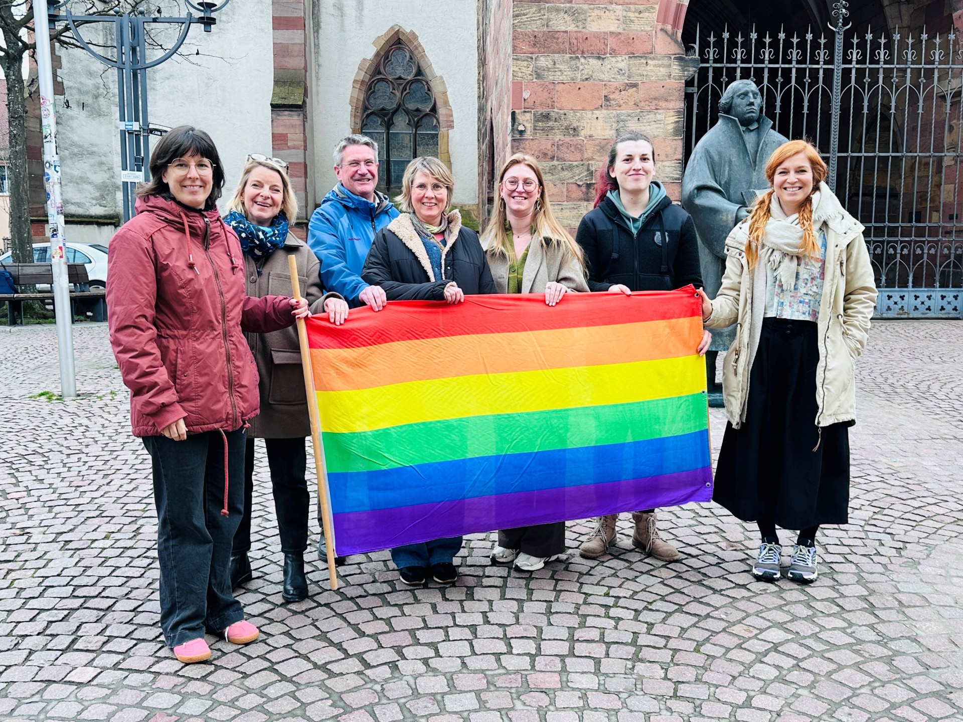 Sieben Personen halten eine Regenbogenflagge und lächeln in die Kamera.