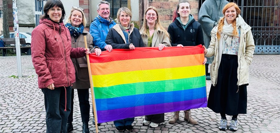 Sieben Personen halten eine Regenbogenflagge und lächeln in die Kamera.