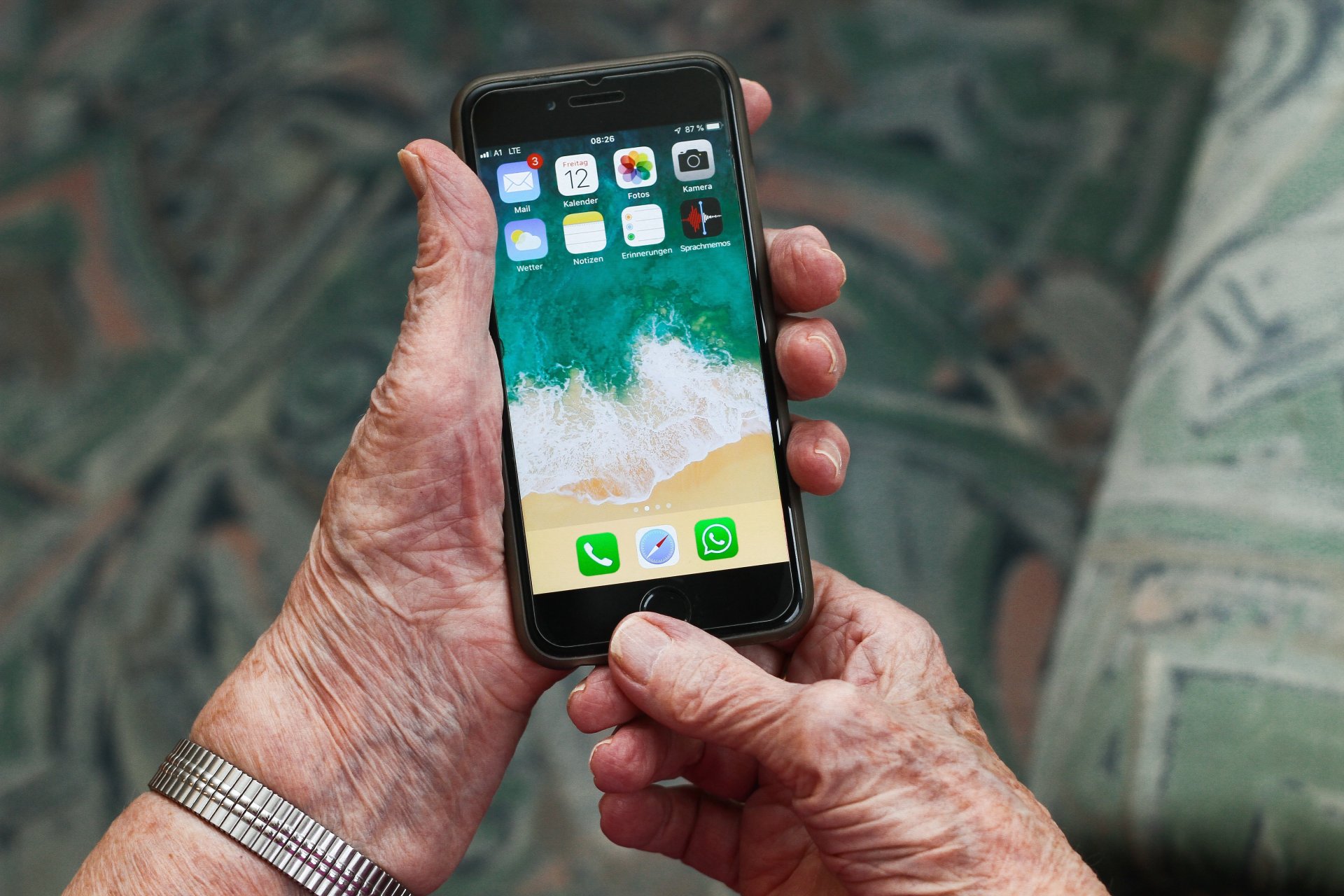 Ältere Person hält ein Smartphone in beiden Händen und bedient den Home-Button. Auf dem Bildschirm sind verschiedene Apps wie E-Mail, Kalender, Kamera und WhatsApp zu sehen. Der Hintergrund ist unscharf.