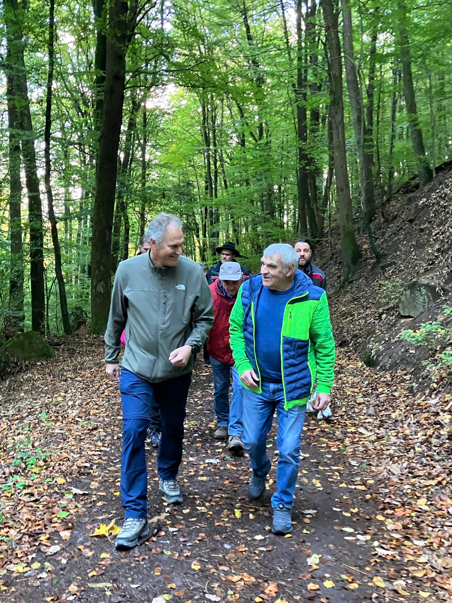 Zwei Männer auf einem Waldweg im Gespräch, im Hintergrund weitere Personen.