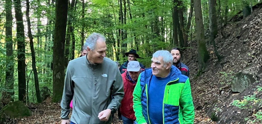 Ernst Keller (rechts) wollte mal mit Landrat Dietmar Seefeldt (links) wandern. Das haben beide im Oktober umgesetzt. Foto: KV SÜW Zwei Männer auf einem Waldweg im Gespräch, im Hintergrund weitere Personen.
