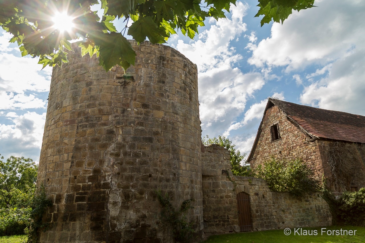 Ein runder Turm aus verwittertem Sandstein, umgeben von historischen mauern. Rechts ist ein steinernes Gebäude mit Satteldach zu sehen. Sonnenstrahlen fallen durch das Laub eines Baumes auf die Anlage, darüber erstreckt sich blauer Himmel mit weißen Wolken.