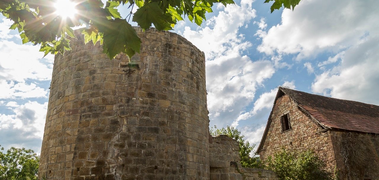 Ein runder Turm aus verwittertem Sandstein, umgeben von historischen mauern. Rechts ist ein steinernes Gebäude mit Satteldach zu sehen. Sonnenstrahlen fallen durch das Laub eines Baumes auf die Anlage, darüber erstreckt sich blauer Himmel mit weißen Wolken.
