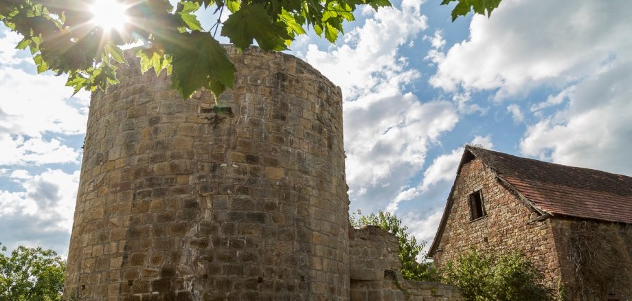 Ein runder Turm aus verwittertem Sandstein, umgeben von historischen mauern. Rechts ist ein steinernes Gebäude mit Satteldach zu sehen. Sonnenstrahlen fallen durch das Laub eines Baumes auf die Anlage, darüber erstreckt sich blauer Himmel mit weißen Wolken.