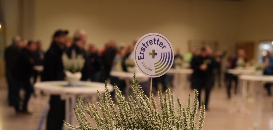 Auf einem Stehtisch steht ein Töpfchen weißes Heidekraut, darin ein Stecker mit der Aufschrift "Erstretter Südpfalz". Im Hintergrund Menschen in Uniform an weiteren Stehtischen.