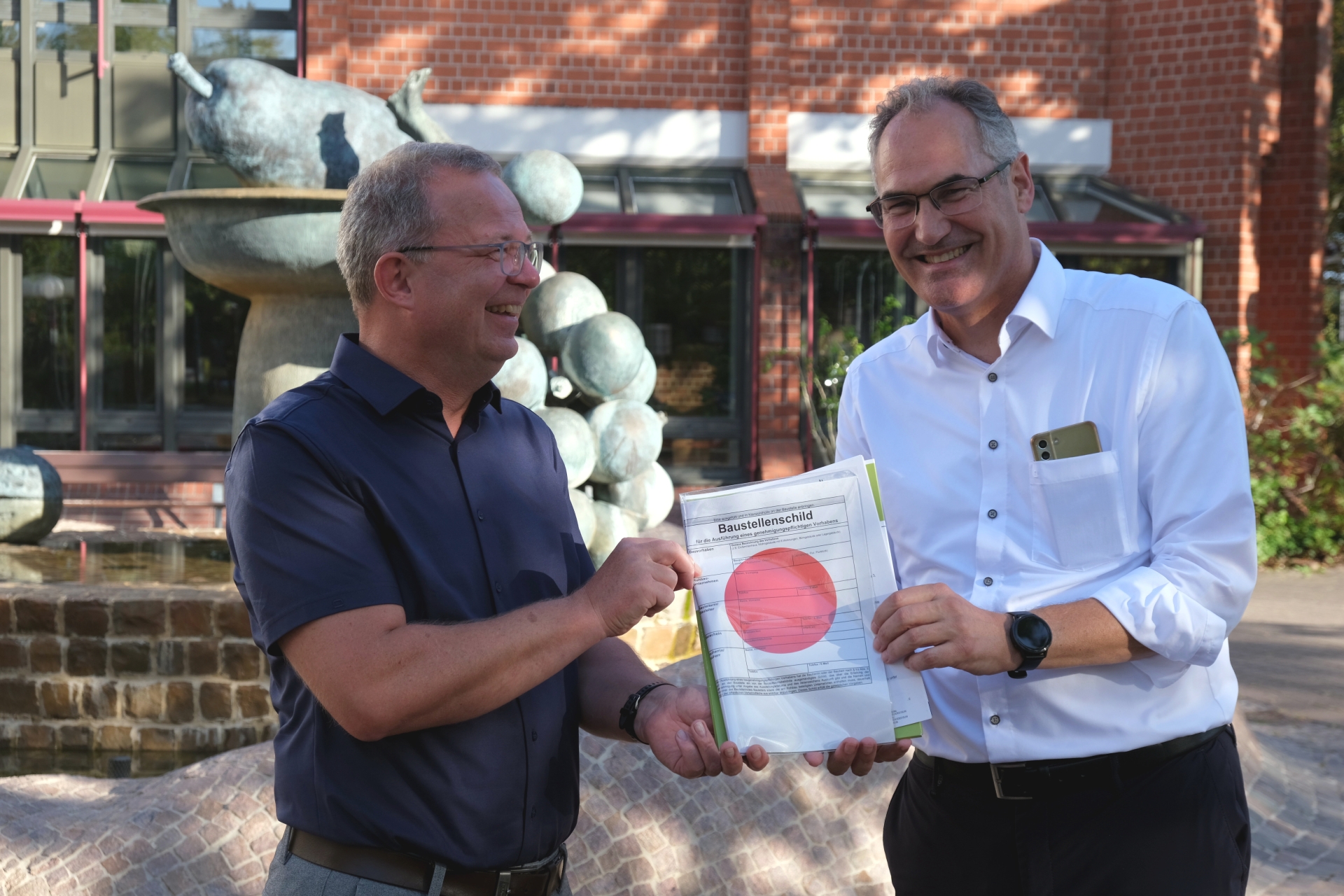Landrat Dietmar Seefeldt übergibt Joachim Freitag vor dem Kreishaus in Landau die Baugenehmigung für die geplante Einrichtung in Offenbach mitsamt dem obligatorischen roten Punkt.