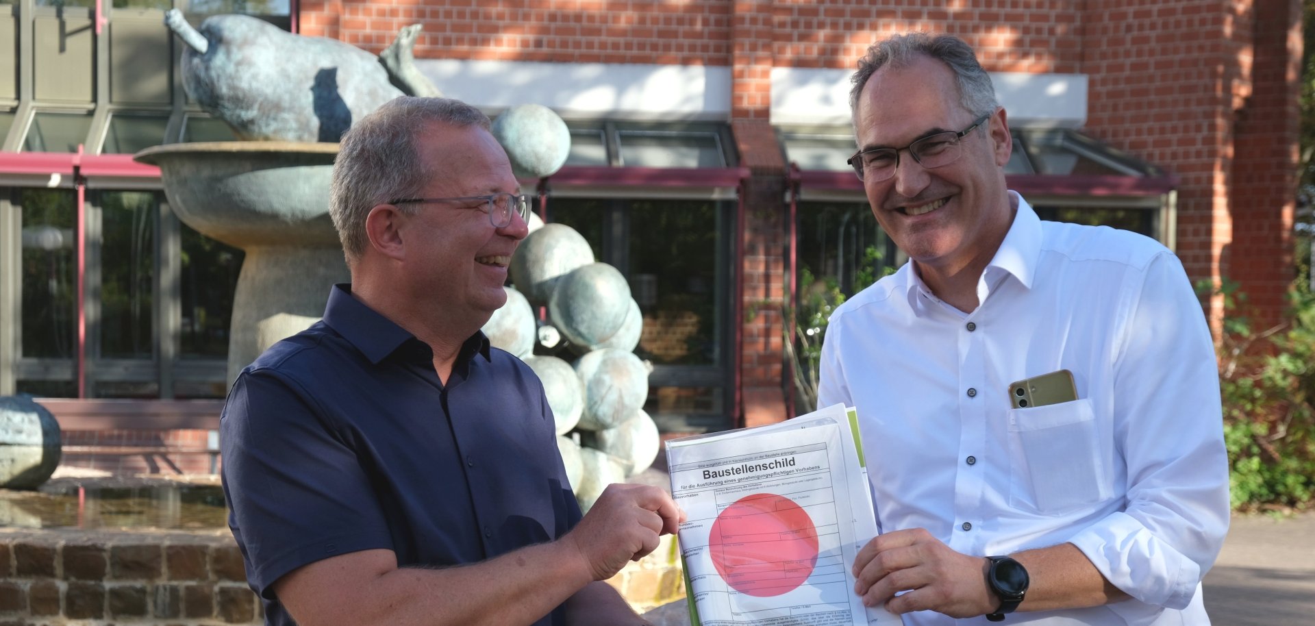 Landrat Dietmar Seefeldt übergibt Joachim Freitag vor dem Kreishaus in Landau die Baugenehmigung für die geplante Einrichtung in Offenbach mitsamt dem obligatorischen roten Punkt.