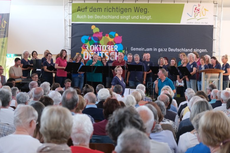 Der Chor „Herztöne“ aus Steinfeld, geleitet von Peter Kusenbach, sowie der Chor „Chorisma“ Insheim unter Leitung von Irina Landes sangen auf der Bühne. Foto: KV SÜW Bühne mit Menschen in bunten T-Shirts, zwei Dirigenten und Publikum.