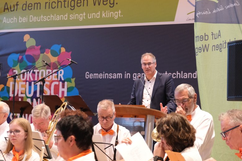 Landrat Dietmar Seefeldt betonte in seiner kurzen Begrüßung, wie wichtig Zusammenhalt sei. Foto: KV SÜW Dietmar Seefeldt auf der Bühne des Dorfgemeinschaftshauses Insheim. Im Hintergrund ein Banner mit Aufschrift en, zu lesen ist: "Auf dem richtigen Weg. Auch bei Deutschland singt und klingt. 3. Oktober. Gemeinsam in ganz Deutschland." Im Vordergrund Musiker mit Blasinstrumenten.