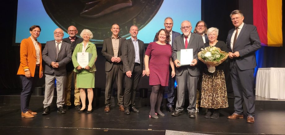 Bildunterschrift: Die stolzen Preisträger Eva Wagner-Seifert aus Klingenmünster (Vierte von links) und Bernd Flaxmeyer aus Birkweiler (Vierter von rechts) sowie Gratulanten von der Südlichen Weinstraße. Foto: KV SÜW Gruppenfoto von elf Personen auf einer Bühne bei einer feierlichen Ehrung.