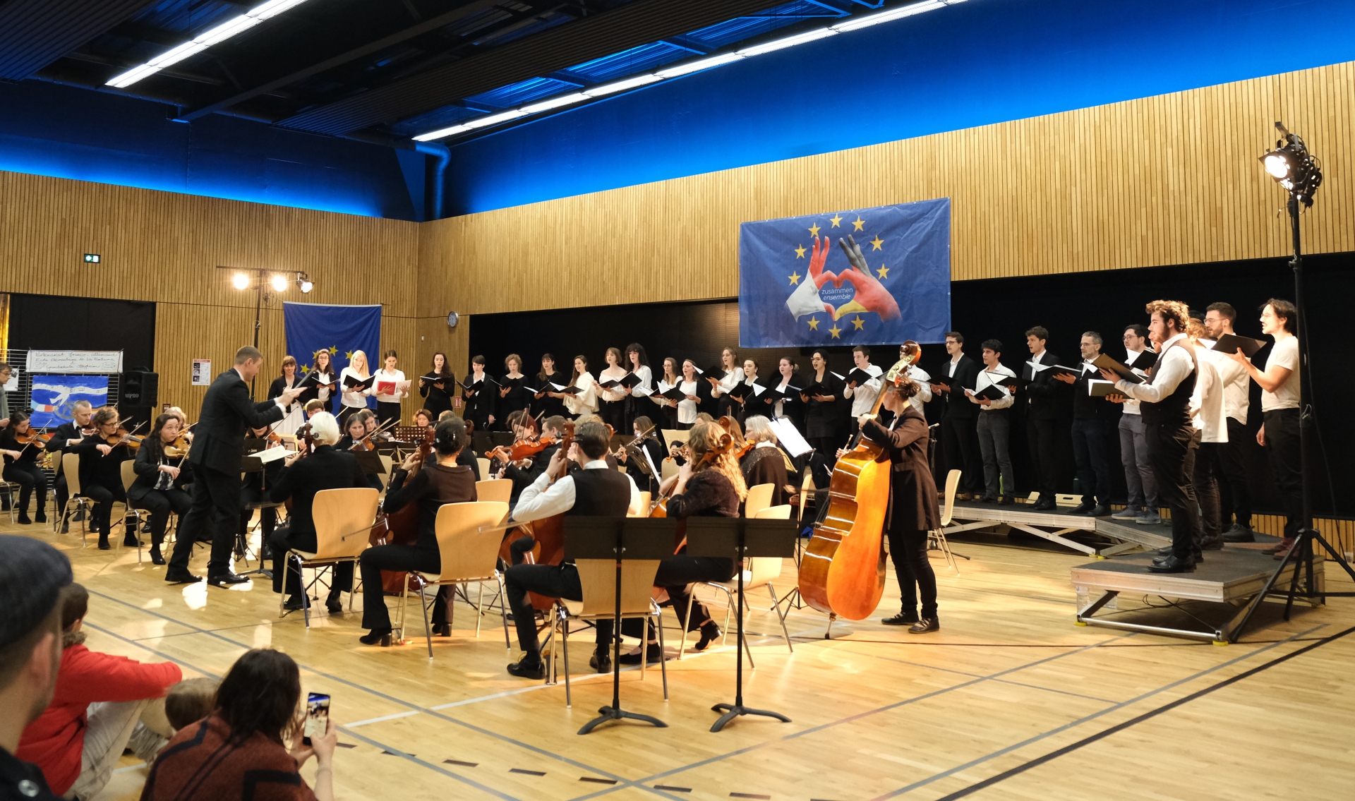Ein Orchester mit Streichinstrumenten und ein gemischter Chor treten gemeinsam in einer Veranstaltungshalle auf. Vorne dirigiert ein Mann die Musikerinnen und Musiker, während der Chor im Hintergrund auf der Bühne singt. An der holzverkleideten Wand hängen blaue Banner mit europäischen Symbolen.