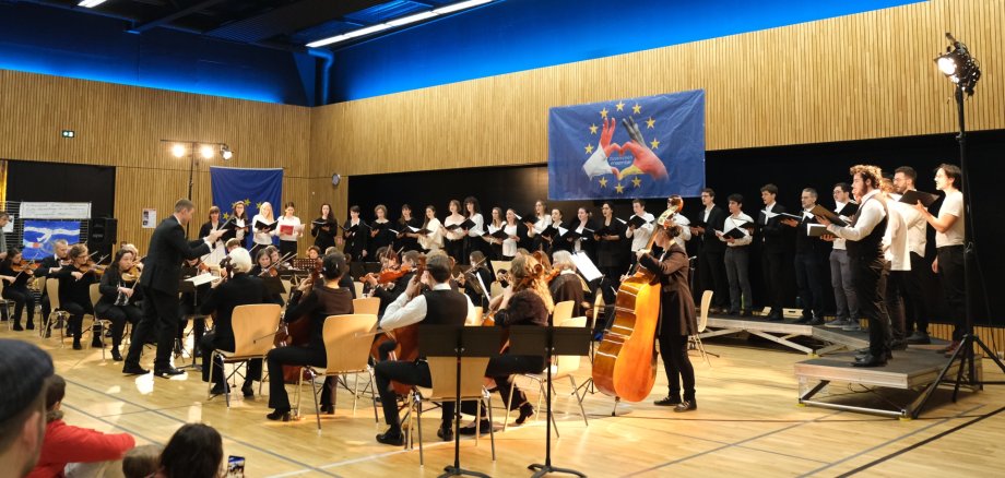 Das deutsch-französische Sinfonieorchester unter Marc Bender – hier im Jahr 2023 zur Feier des 60-jährigen Bestehens des Elysée-Vertrags – wird diesen November wieder auftreten. Archivfoto: KV SÜW Ein Orchester mit Streichinstrumenten und ein gemischter Chor treten gemeinsam in einer Veranstaltungshalle auf. Vorne dirigiert ein Mann die Musikerinnen und Musiker, während der Chor im Hintergrund auf der Bühne singt. An der holzverkleideten Wand hängen blaue Banner mit europäischen Symbolen.