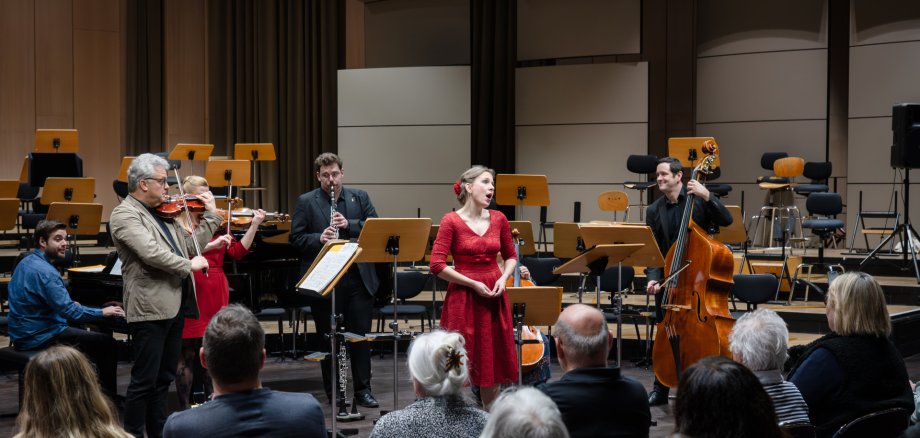 Bei den demenzsensiblen Konzerten mit der Deutschen Staatsphilharmonie Rheinland-Pfalz sind Menschen mit Demenz und ihre pflegenden Angehörigen eingeladen, gemeinsam einen musikalischen Nachmittag zu erleben. (Quelle: Christian Kleine) Eine Sängerin inmitten eines Ensembles an Musikern, im Vordergrund Publikum.