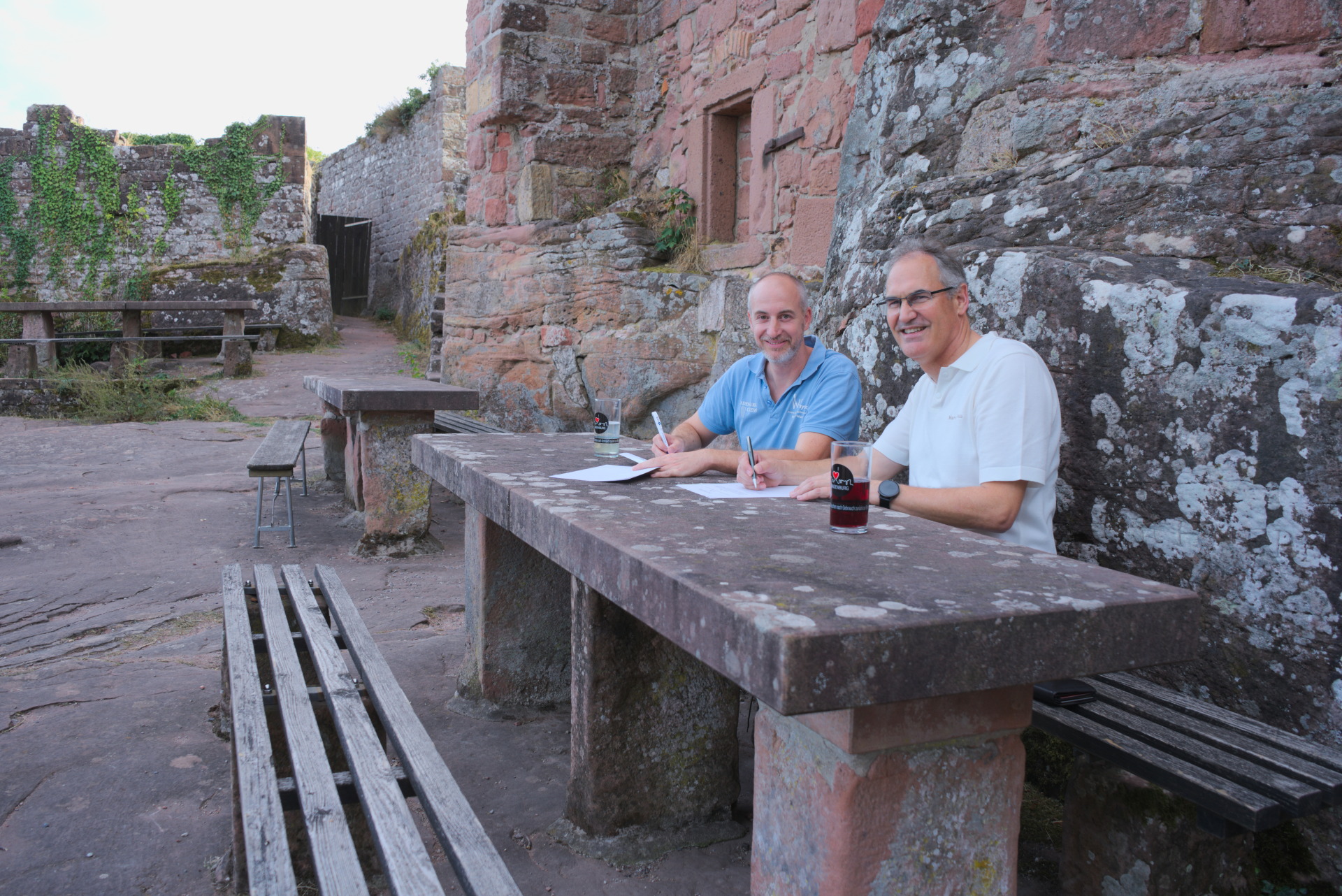 Zwei Männer sitzen lächelnd an einem massiven Steintisch inmitten einer historischen Burgruine aus rötlichem Sandstein. Vor ihnen liegen Unterlagen, auf denen sie mit Stiften schreiben.