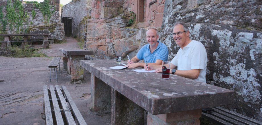 Landrat Dietmar Seefeldt (rechts), Vorsitzender des Madenburgvereins, und Pächter Sven Buchwald bei der Unterzeichnung des neuen Pachtvertrags für die Madenburgschänke. Foto: KV SÜW Zwei Männer sitzen lächelnd an einem massiven Steintisch inmitten einer historischen Burgruine aus rötlichem Sandstein. Vor ihnen liegen Unterlagen, auf denen sie mit Stiften schreiben.