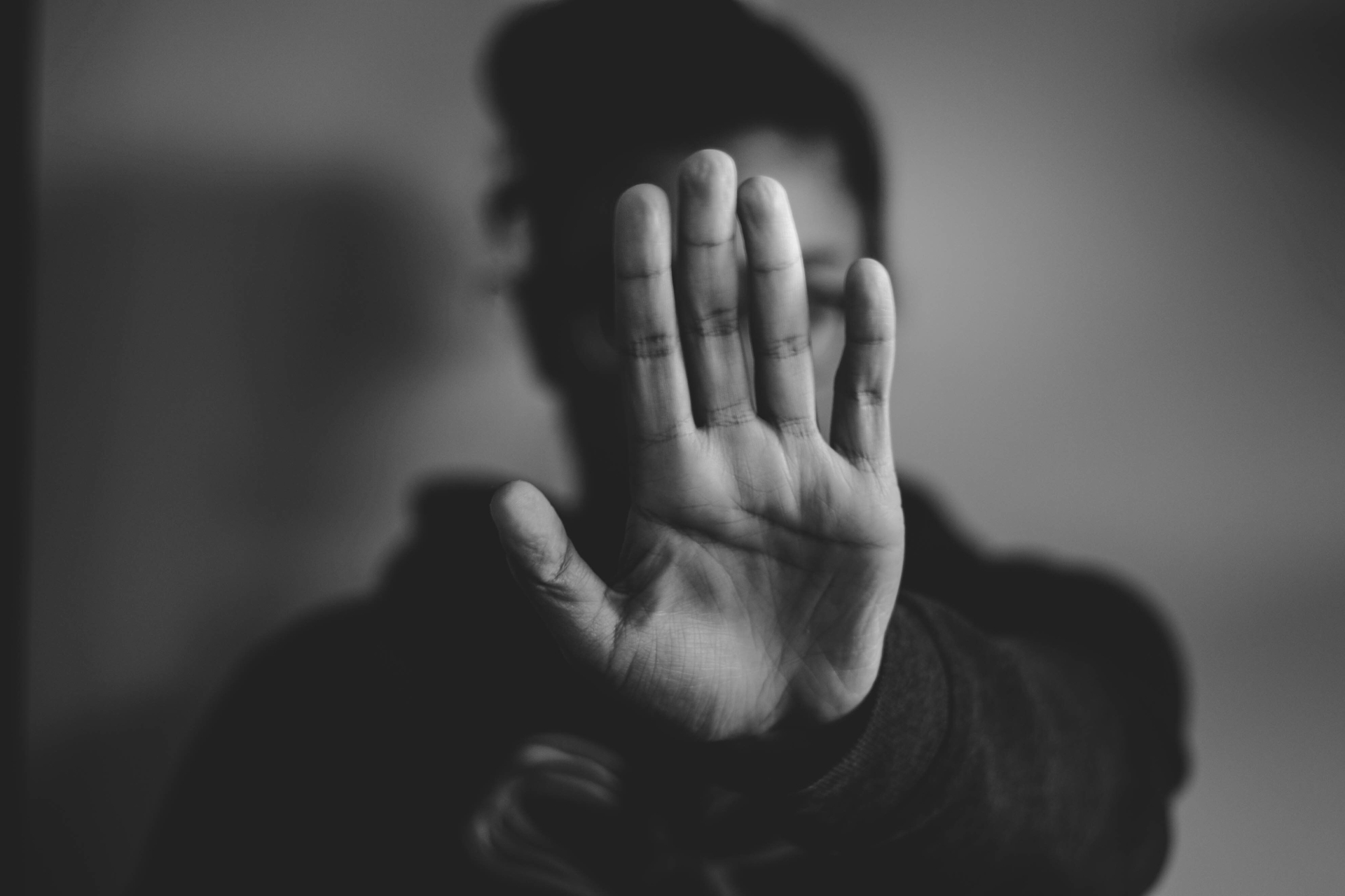 Schwarz-weiß-Fotografie einer Person, die ihre Hand mit der Handfläche nach vorne abwehrend vor ihr Gesicht hält.