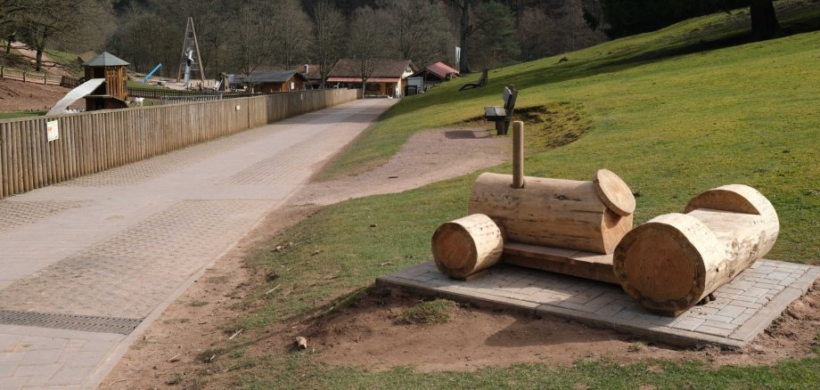 Spielmöglichkeiten wie dieser Traktor aus Holz locken entlang der Wanderwege im Wild- und Wanderpark Südliche Weinstraße. Foto: KV SÜW Im Vordergrund ein Traktor aus Holz, im Hintergrund eine Riesenrutsche im Wildpark Silz.