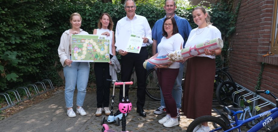 Landrat Dietmar Seefeldt (Mitte) überbrachte Urkunde und Spielteppich an die kommunale Kita in Hochstadt, hier vertreten durch die Leitung Claudia Bechtel (vorne links) und stellvertretende Leitung Celine Kaifer (vorne rechts) sowie Ortsbürgermeister Timo Reuther. SÜW-Klimaschutzmanagerin Agnes Bartmus (links) und Pia Werling, Beigeordnete der Verbandsgemeinde Offenbach an der Queich (2. von links), zeigen einen exemplarischen Spielplan der Aktion. Foto: KV SÜW Eine Gruppe von sechs Personen steht im Freien vor einer begrünten Wand. Zwei Personen halten Plakate in der Hand, zwei weitere zusammengerollte Stoffbahnen.