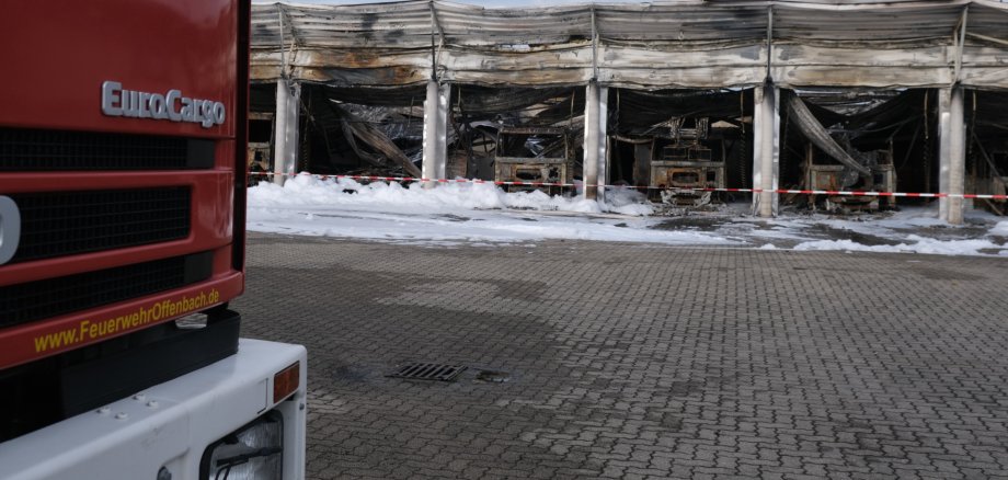 Im Vordergrund ein Feuerwehrfahrzeug, im Hintergrund eine ausgebrannte Halle und weißer Schaum.