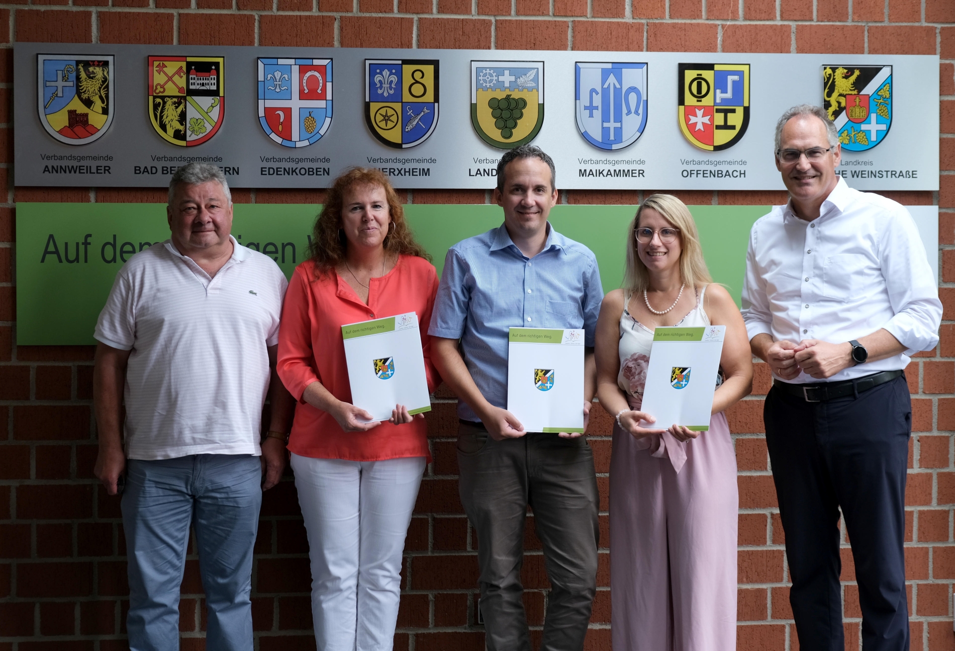 Fünf Personen stehen nebeneinander vor einer Wand mit den Wappen des Landkreises Südliche Weinstraße und er zugehörigen  Verbandsgemeinden. Drei von ihnen halten Urkunden in den Händen.
