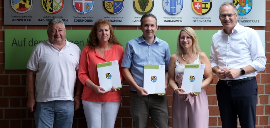 Landrat Dietmar Seefeldt (rechts) und Personalratsvorsitzender Udo Baumgartner (links) mit den Jubilarinnen Edith Reinhard (2. von links) und Jennifer Pfaffmann (2. von rechts) sowie dem Jubilar Joscha Wittiber (Mitte). Foto: KV SÜW Fünf Personen stehen nebeneinander vor einer Wand mit den Wappen des Landkreises Südliche Weinstraße und er zugehörigen Verbandsgemeinden. Drei von ihnen halten Urkunden in den Händen.
