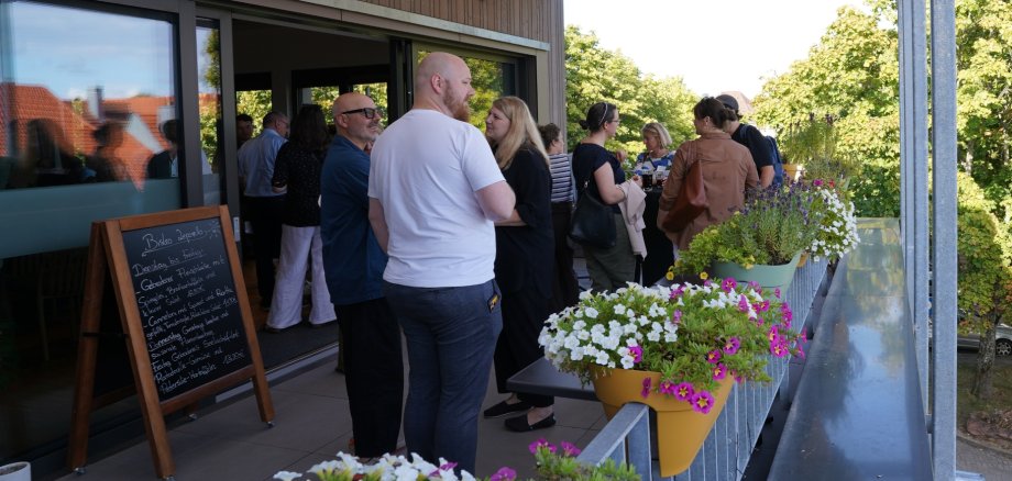 Mehrere Personen stehen bei Sonnenschein auf einer Terrasse zusammen und unterhalten sich.