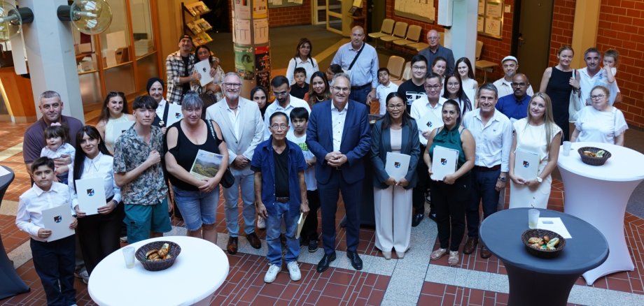 Nach der feierlichen Einbürgerungsfeier im Kreishaus SÜW. Foto: KV SÜW Eine größere Gruppe von Erwachsenen und Kindern steht für ein Gruppenfoto im Foyer der Kreisverwaltung. Mehrere Personen halten Urkunden in den Händen.