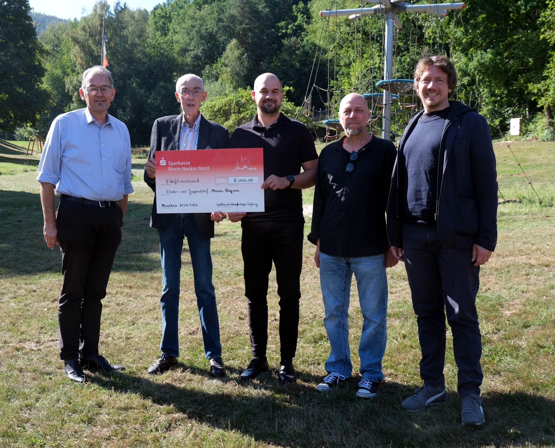Fünf Männer stehen auf einer Wiese vor einem Kletterparcours und posieren für ein Gruppenfoto. Zwei von ihnen halten einen symbolischen Spendenscheck über 5000 Euro in den Händen.