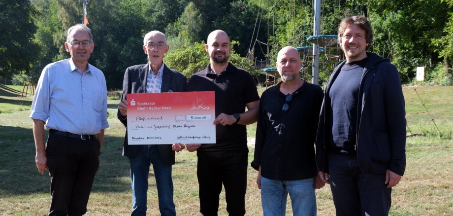 Fünf Männer stehen auf einer Wiese vor einem Kletterparcours und posieren für ein Gruppenfoto. Zwei von ihnen halten einen symbolischen Spendenscheck über 5000 Euro in den Händen.