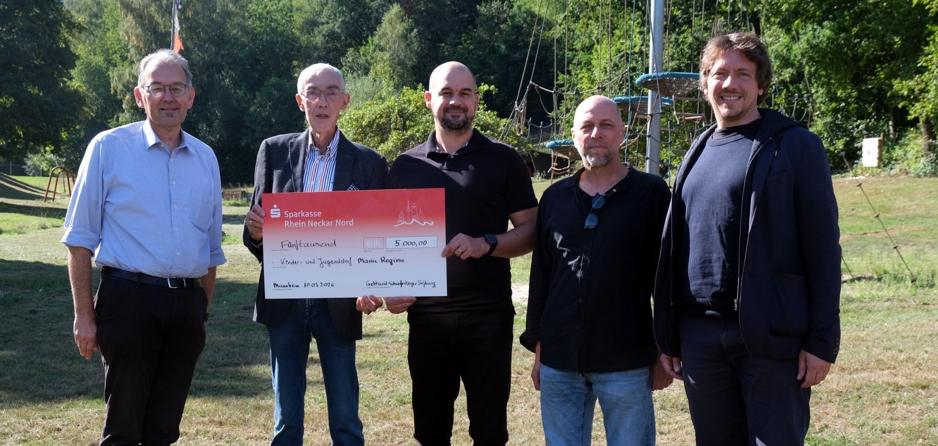 Fünf Männer stehen auf einer Wiese vor einem Kletterparcours und posieren für ein Gruppenfoto. Zwei von ihnen halten einen symbolischen Spendenscheck über 5000 Euro in den Händen.