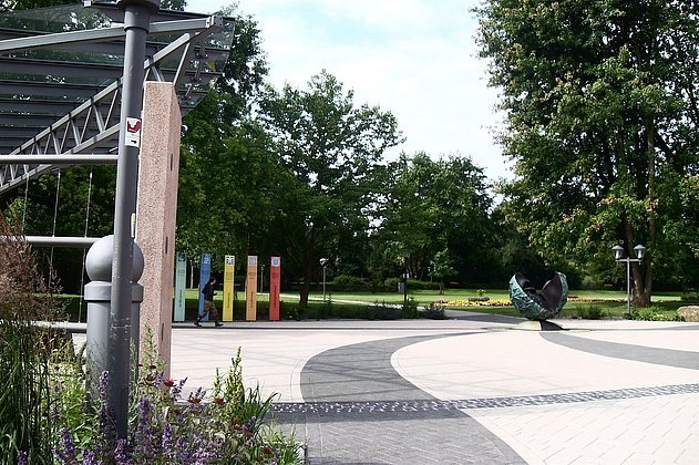Blick auf einen modern gestalteten Platz mit geschwungenen Pflasterflächen, bunten Informationsstelen und einer Skulptur. Bäume und Grünflächen erstrecken sich dahinter.